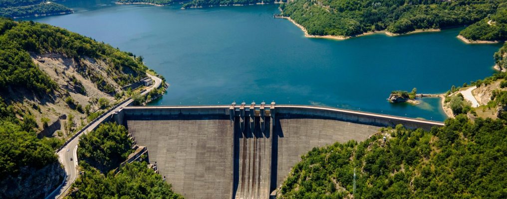 Vista aérea de uma barragem hidrelétrica cercada por montanhas cobertas de vegetação e um grande reservatório de água azul, com uma estrada sinuosa à esquerda e uma pequena ilha à direita.